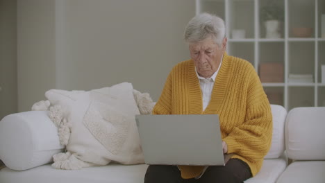 An-elderly-woman-sitting-on-the-couch-prints-a-message-on-the-keyboard.-Grandma-uses-a-laptop